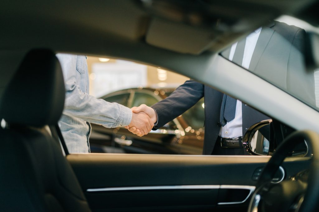 Buyer of car shaking hands with seller in auto dealership, view from interior of car. Close-up of handshake of business people. Concept of choosing and buying new car at showroom.