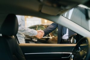 Buyer of car shaking hands with seller in auto dealership, view from interior of car. Close-up of handshake of business people. Concept of choosing and buying new car at showroom.